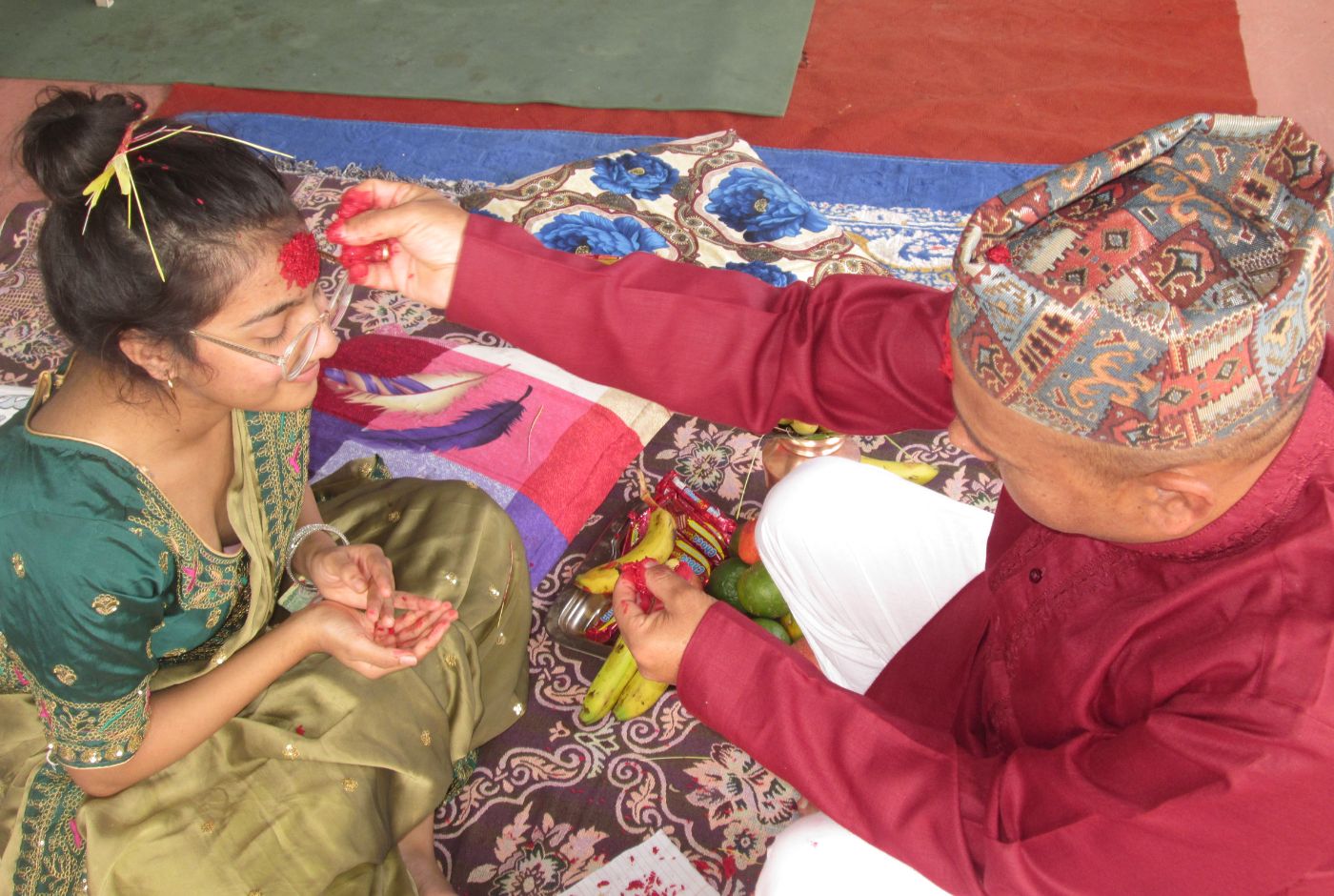Elderly man places red tika and jamara on a young woman&rsquo;s forehead during Dashain Festival, as she sits respectfully to receive blessings.