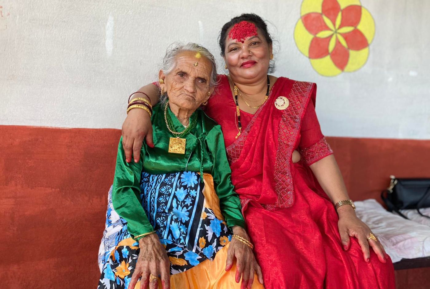 Elderly Nepali woman and younger woman sitting together during Dashain, wearing traditional dress with red tika on their foreheads.