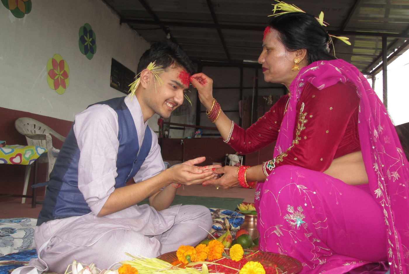 A woman places red tika and jamara on a young man&rsquo;s forehead during Dashain Festival, as he smiles and receives blessings seated indoors.