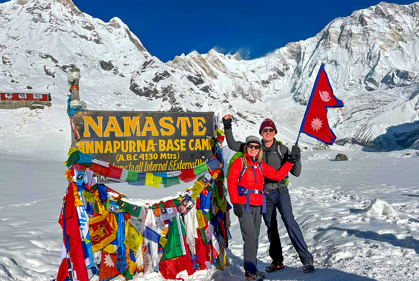 Two trekkers celebrating triumphantly at Annapurna Base Camp sign decorated with colorful prayer flags and Nepal flag, snow-covered Annapurna massif and glacier in background under clear blue sky, marking successful completion with proper ACAP permits