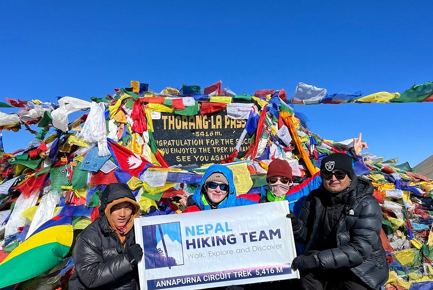 Trekkers celebrating at Thorong La Pass (5,416m) with prayer flags and Nepal Hiking Team banner on Annapurna Circuit Trek