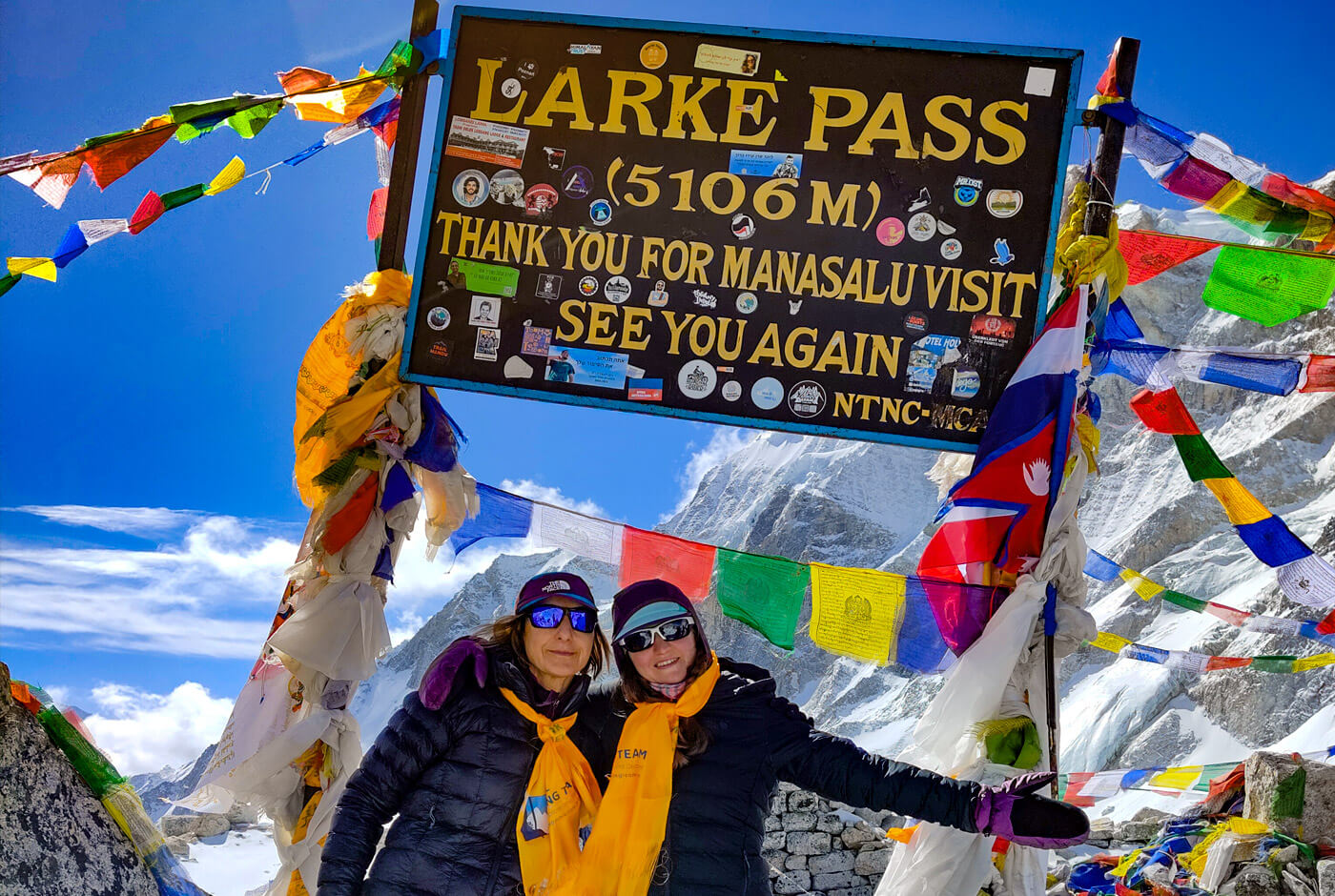 Two female trekkers at Larkya La Pass 5160m sign with prayer flags on the Manaslu Circuit Trek Nepal