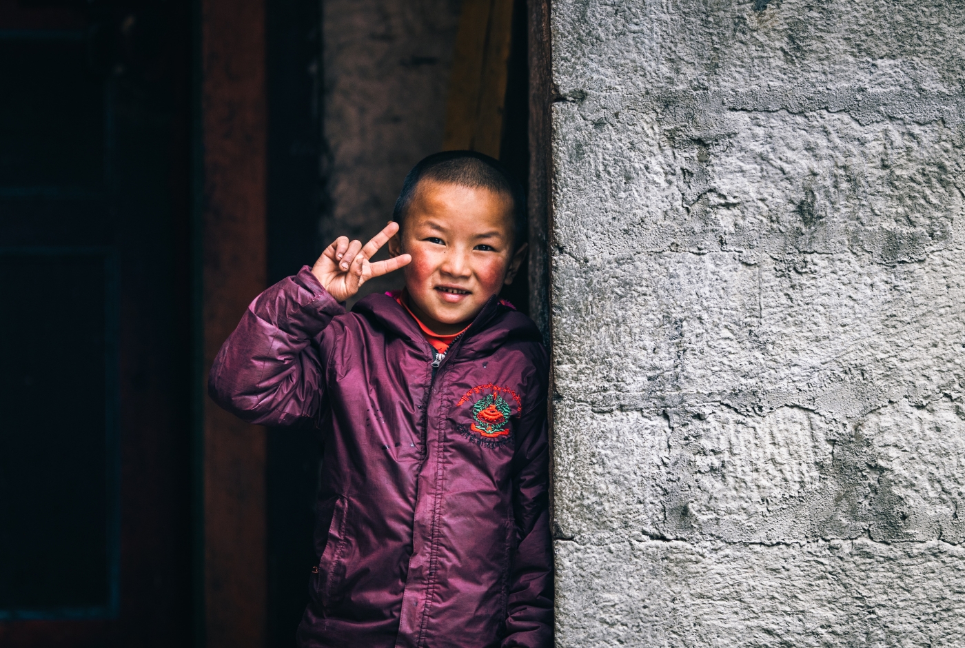 A smiling child in a purple jacket stands by a stone wall, making a peace sign   Thame village